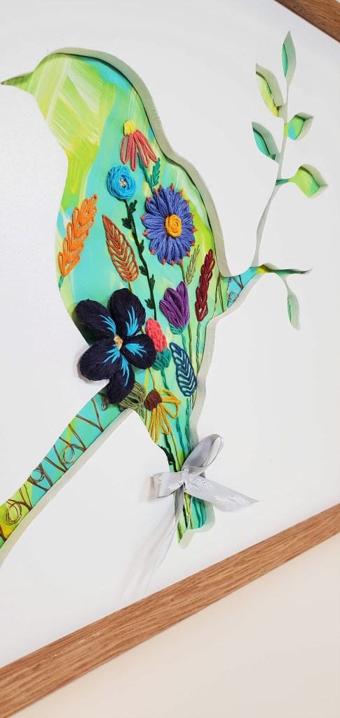 Close up detail of a bird sitting on branch silhouette with a blue background and embroidered flower details.