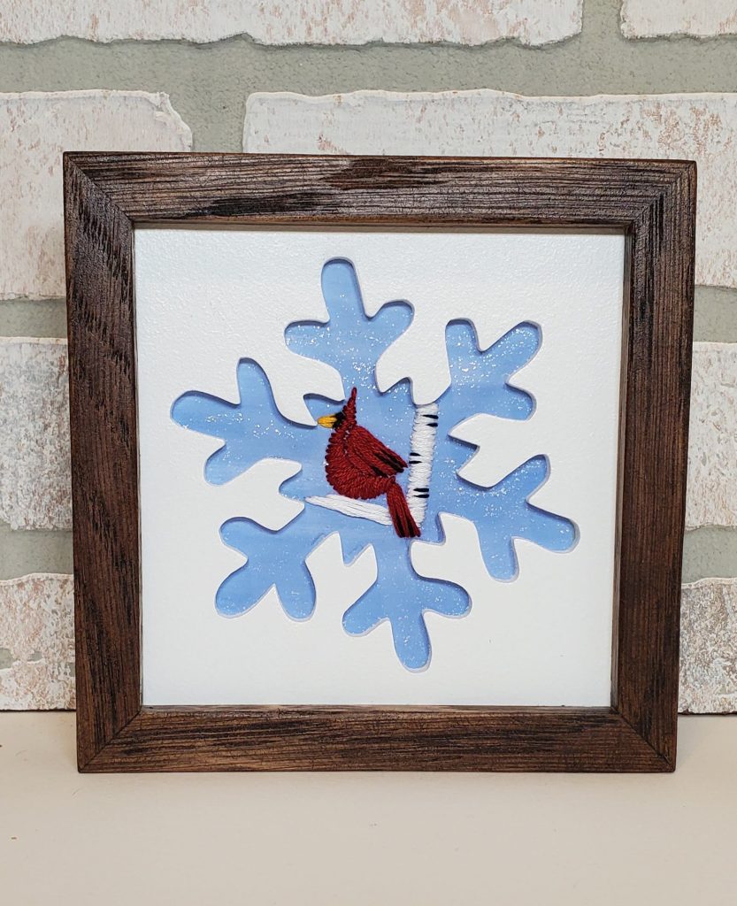 Snowflake silhouette with blue background and embroidered cardinal sitting on a birch branch.