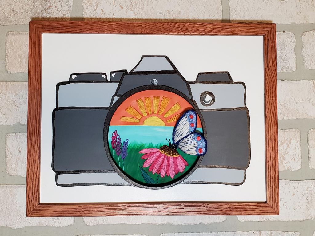 Vintage looking camera with a sunset background inside the lens with an embroidered butterfly sitting on a flower detail.