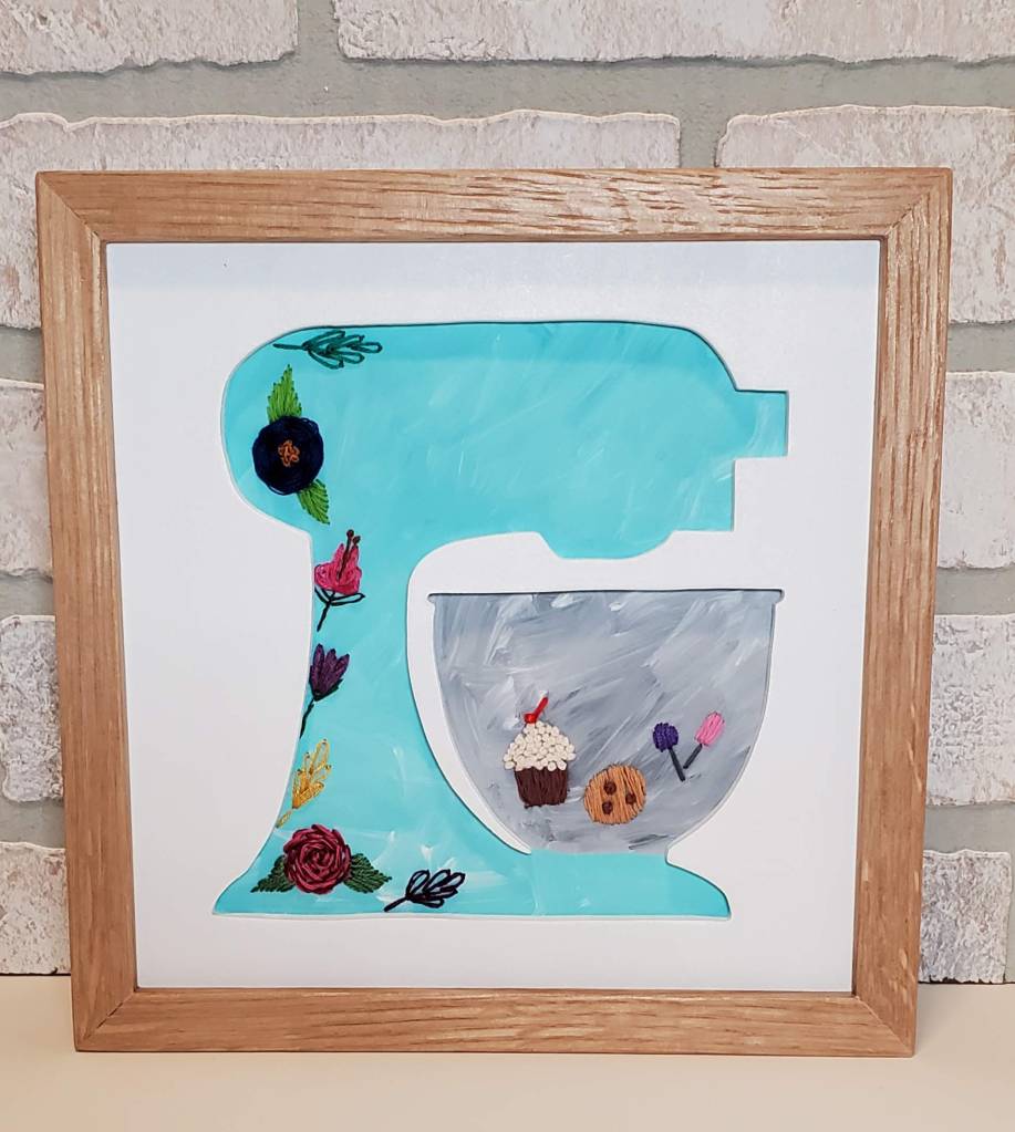 Stand-up baking mixer silhouette with teal background, embroidered floral designs, cupcake and cookie embroidery inside of mixing bowl.