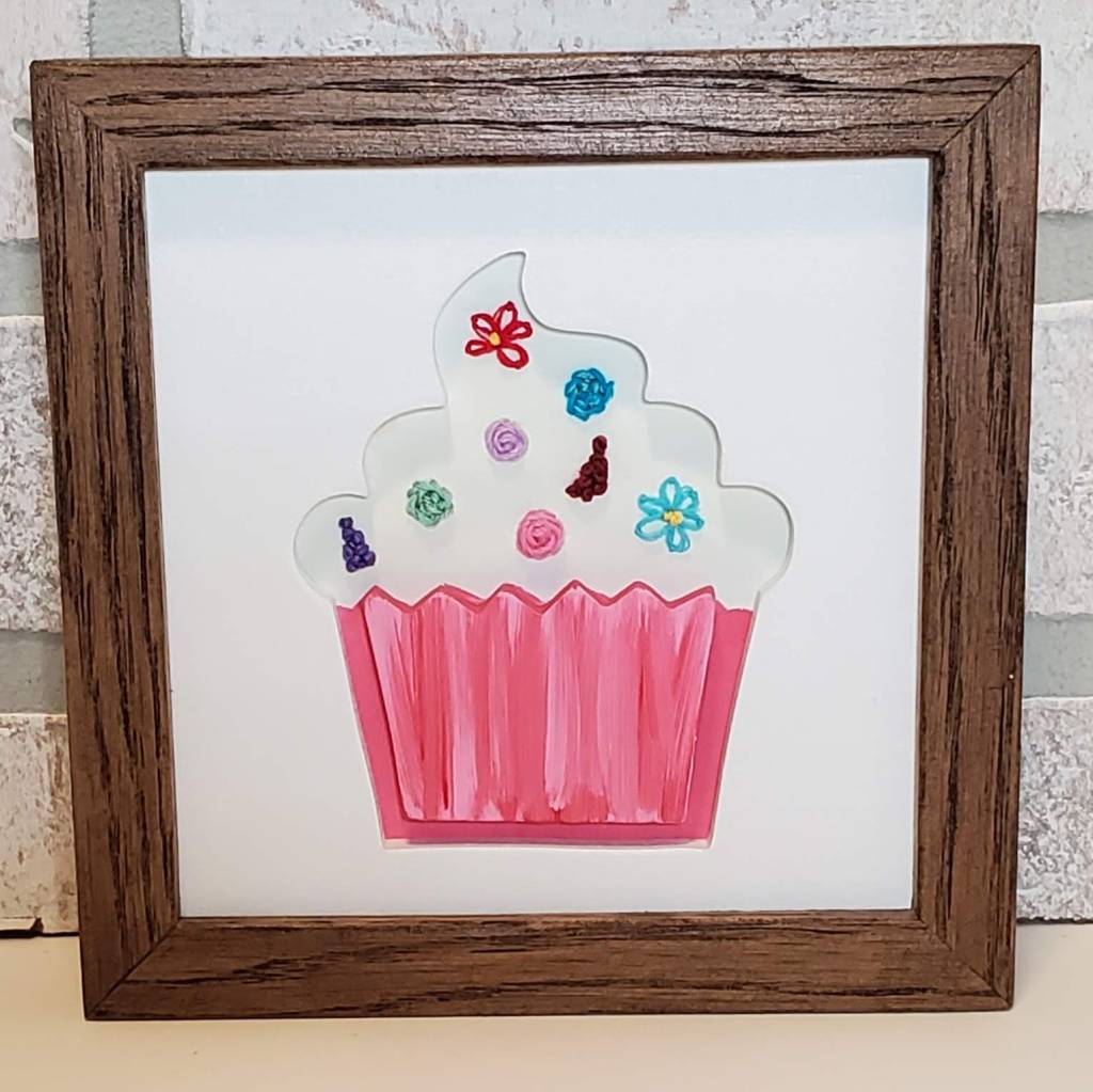Cupcake silhouette with pink bottom and white frosting. Embroidered floral details in the frosting.