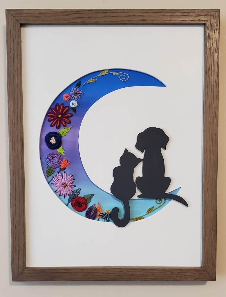 Crescent moon silhouette with blue background and embroidered floral details. A black cat and dog sit in the crescent of the moon.