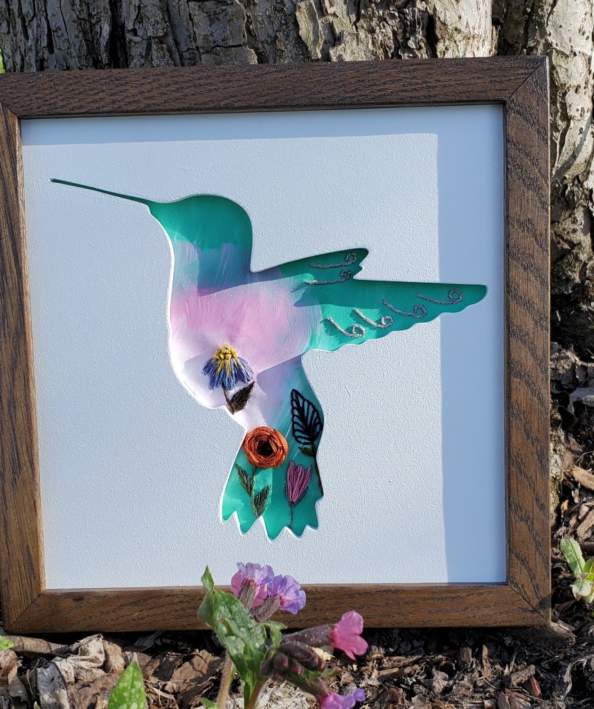 Hummingbird side view silhouette with pastel green and pink background with embroidered flower details.
